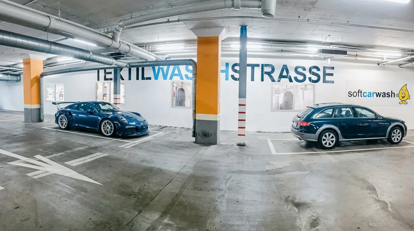Zwei Autos, die in einer Tiefgarage in der Nähe einer Wand mit der Aufschrift „Textilwaschstrasse“ geparkt sind. Zwei Autos, die in einer Tiefgarage in der Nähe einer Wand mit der Aufschrift „Textilwaschstrasse“ geparkt sind.